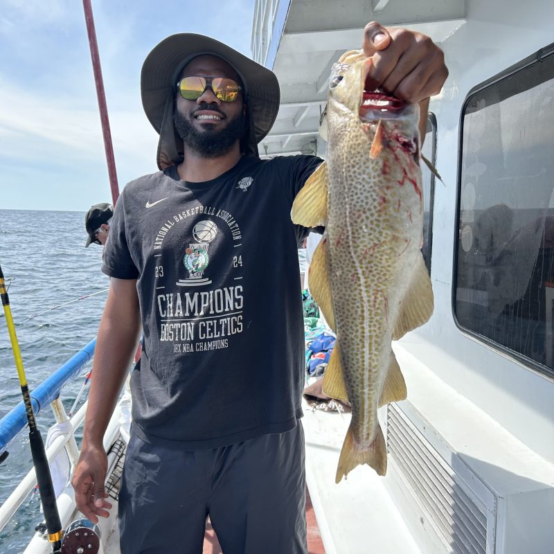 Man on boat holding a large fish, wearing sunglasses and a Boston Celtics shirt.