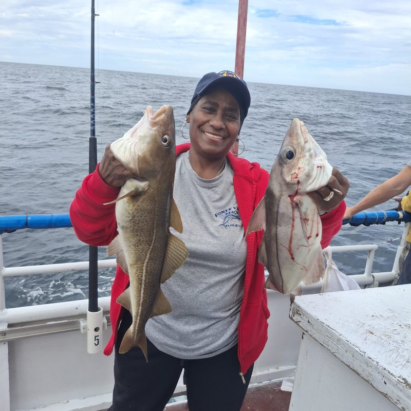 Person smiling on a boat holding two large fish, wearing a cap and red jacket.