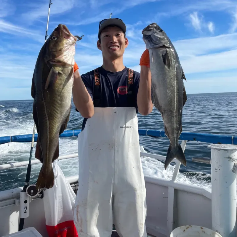 Person on a boat holding two large fish, wearing a cap and white overalls against a blue sky.