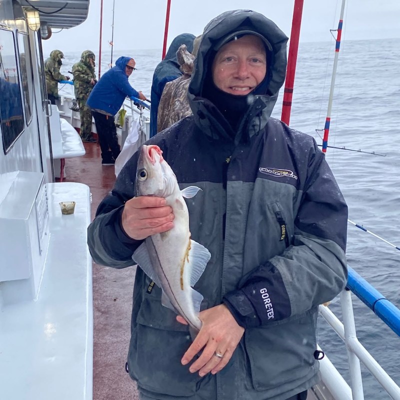 Person in winter gear holding a fish on a boat with others fishing nearby.