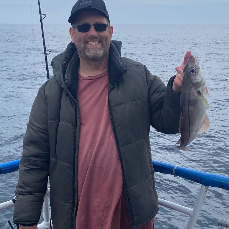Man in jacket and cap holding a fish on a boat at sea.