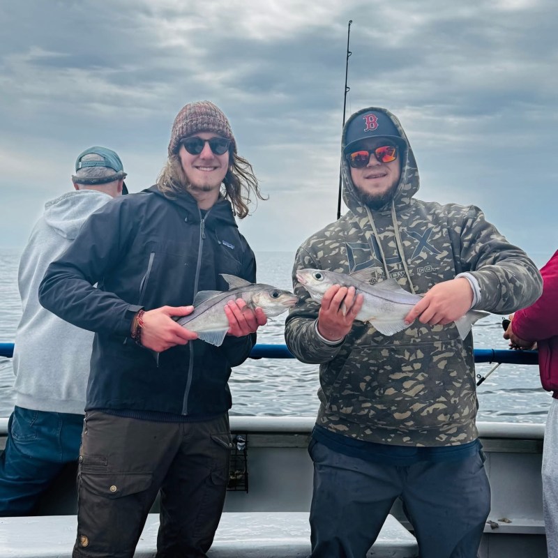 Two people on a boat holding fish against a cloudy sky.