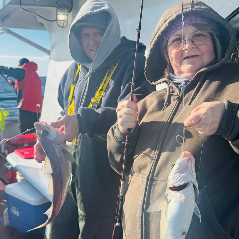 Two people on a boat holding fish, dressed in hoodies and fishing gear.