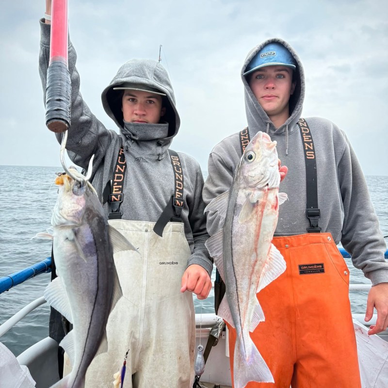 Two people in hoodies and coveralls holding fish on a boat.