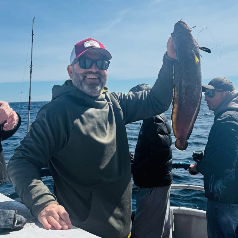 Man on boat proudly holding a fish, wearing a cap and sunglasses.