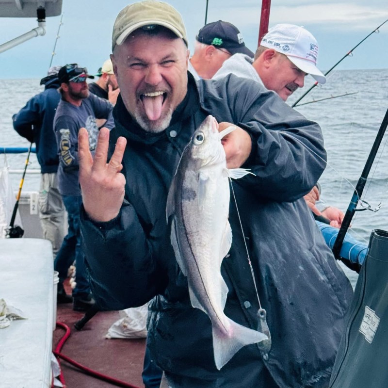 Man on boat holding fish, smiling and gesturing with tongue out.