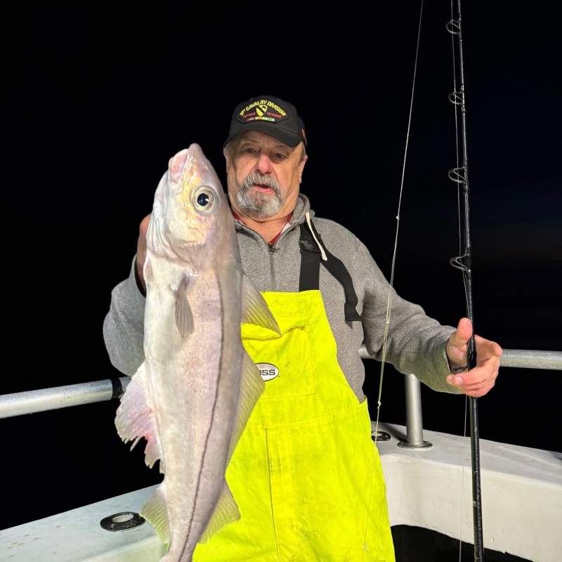 Man in bright yellow overalls holding a large fish on a boat at night.