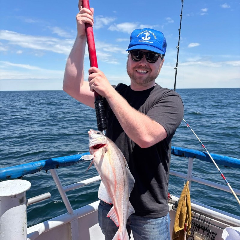 Man smiling, wearing blue cap, holding a fish on a pole on a boat, ocean in the background.