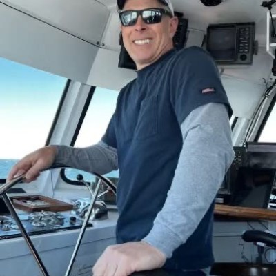 Man smiling at boat helm, wearing sunglasses, cap, and layered shirts.