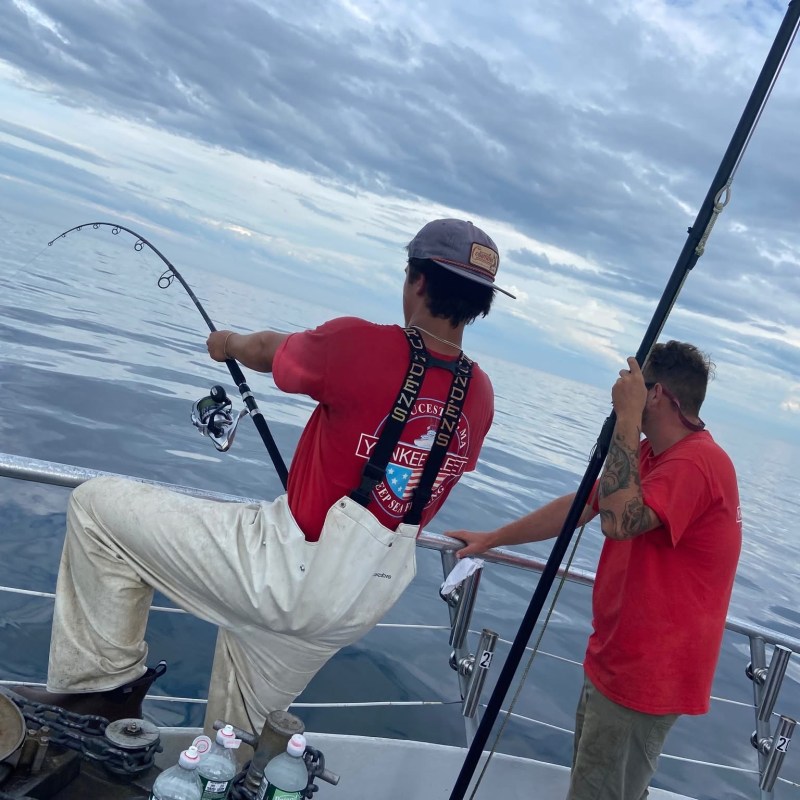 Two people fishing on a boat, one holding a fishing rod, under a cloudy sky.