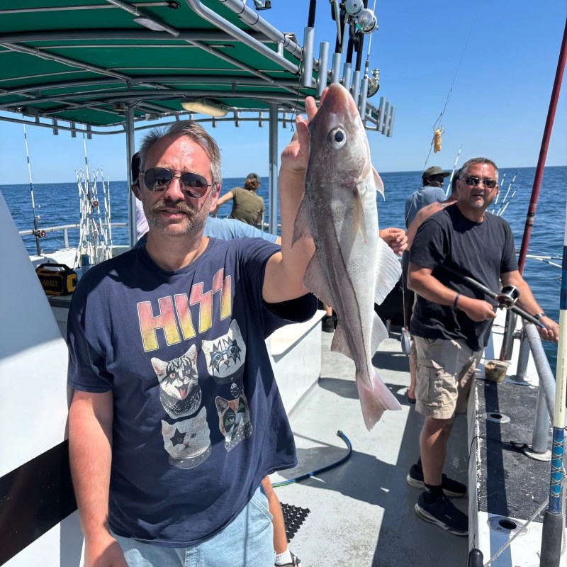Man on boat holding fish, wearing graphic tee, with ocean background.