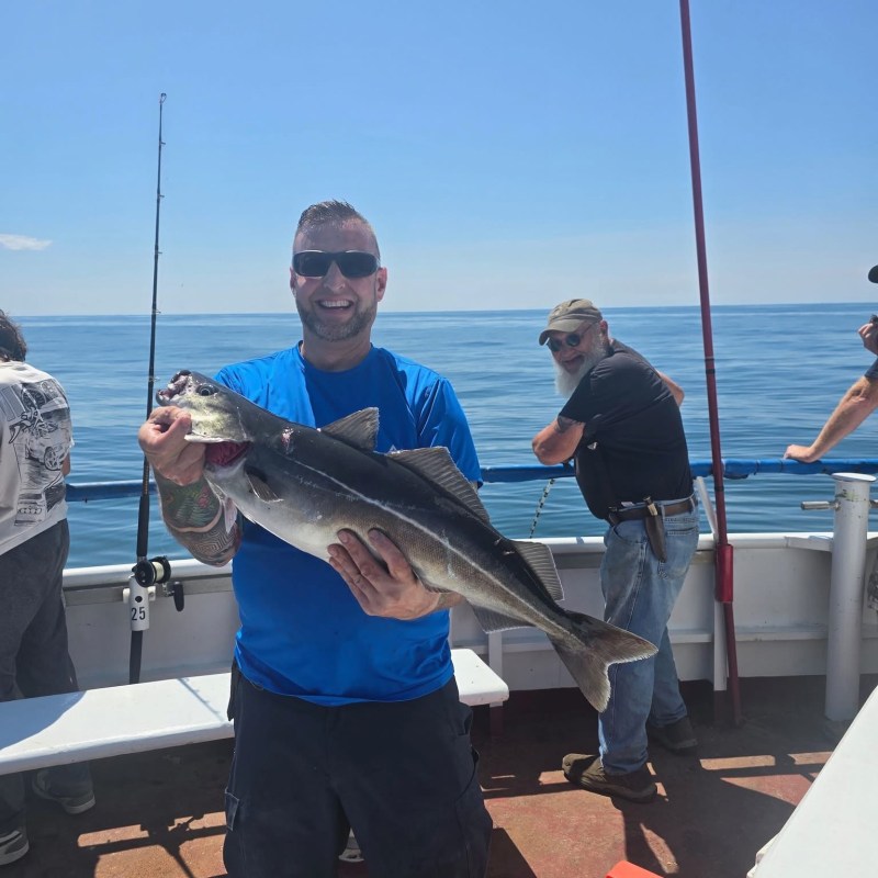 Man on boat holding a large fish, ocean in background, sunny day.