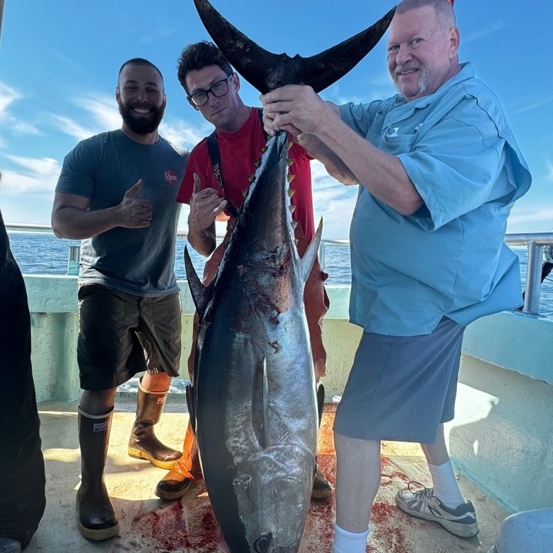 Three men posing with a large tuna on a boat, two holding it upright.