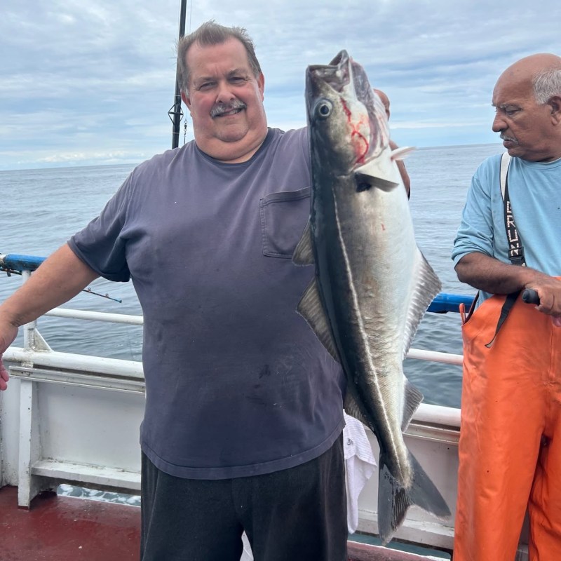 Man on a boat holding a large fish, another man looks on, with cloudy sky and ocean in the background.