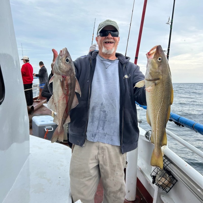 Man on a boat holding two large fish, smiling, with ocean in the background.